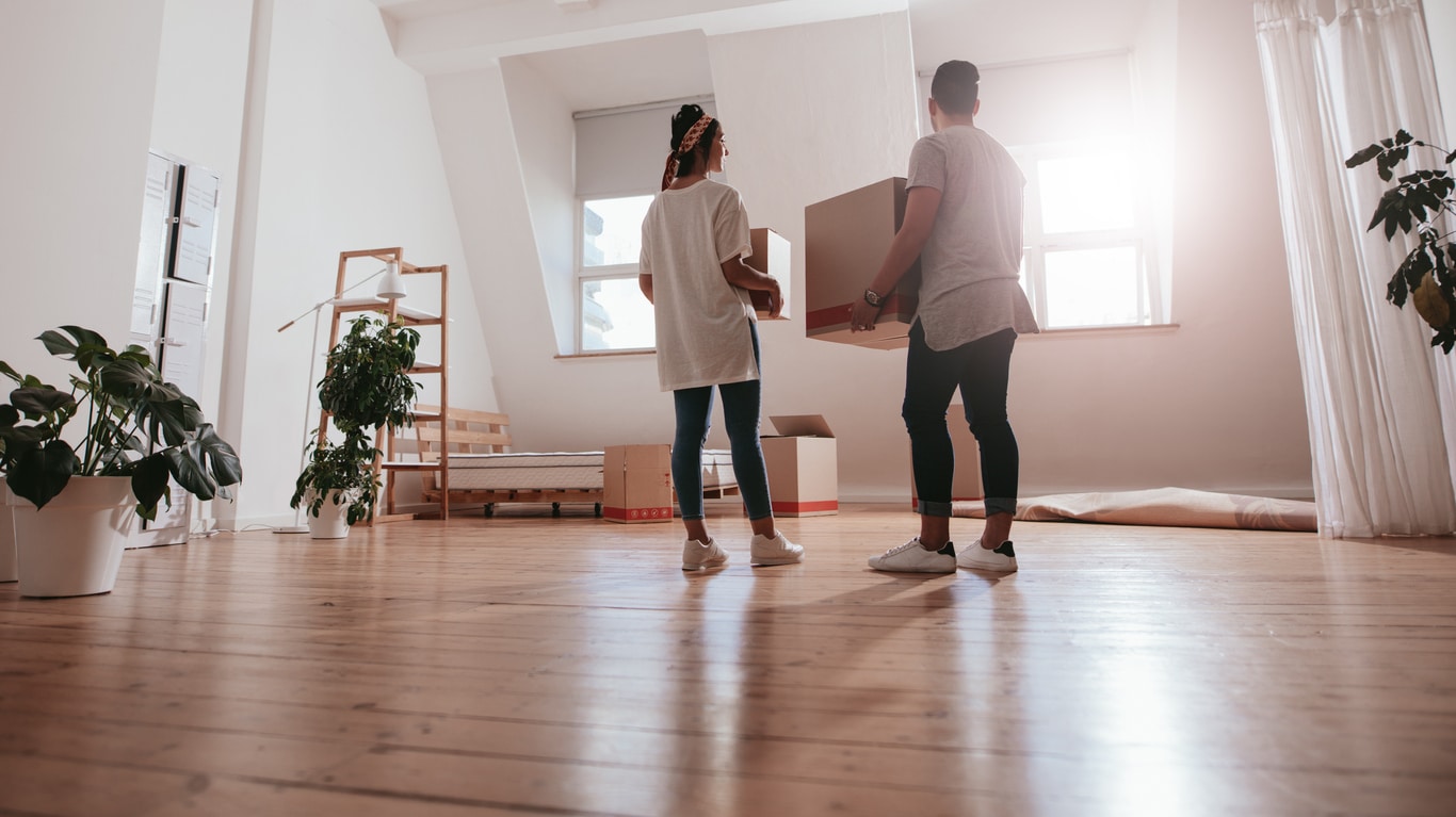 A couple in white t-shirts holding moving boxes pause in their new apartment after moving from a house to an apartment