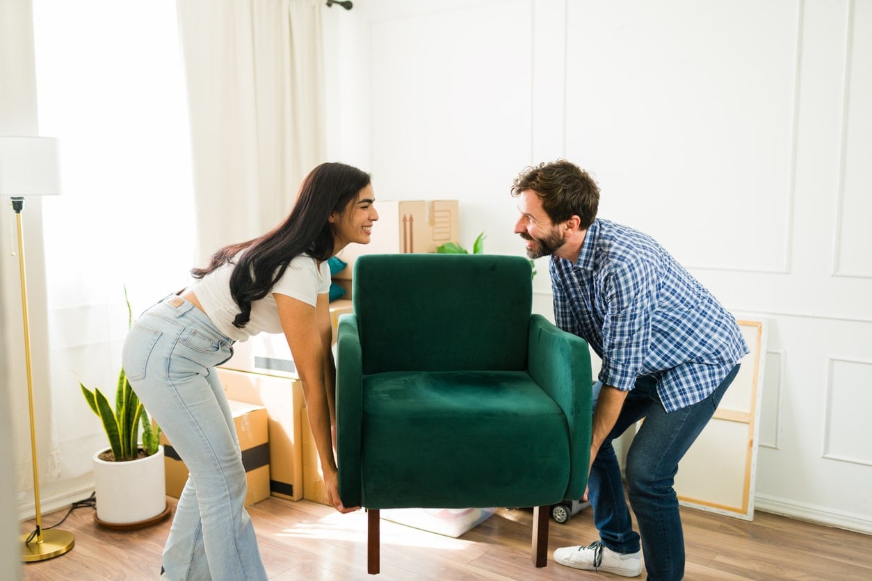 A smiling man and woman carry a green velvet armchair into a bright living room with moving boxes