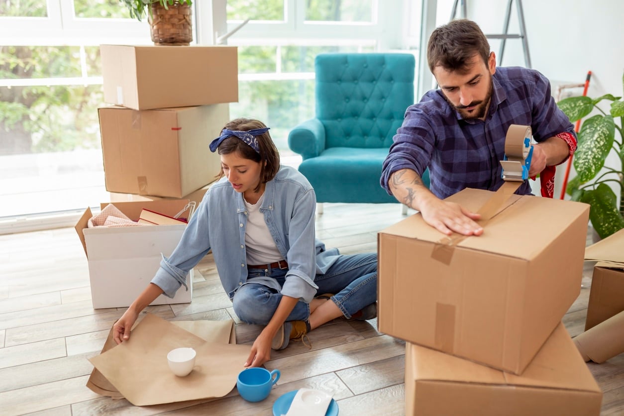 A couple getting ready for their DIY in their living room, with the man using a tape dispenser to seal a moving box
