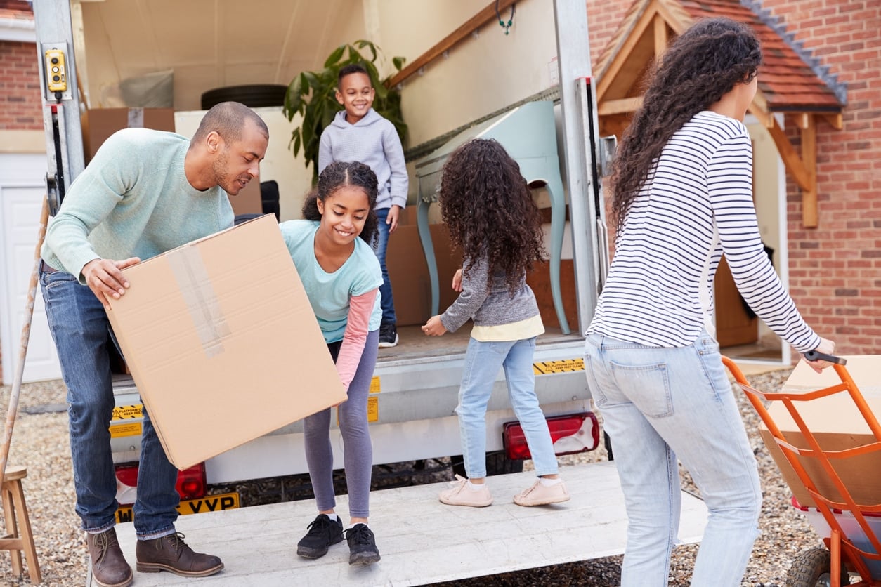 A family in the middle of DIY moving unloads cardboard boxes from the back of a moving truck into their new home
