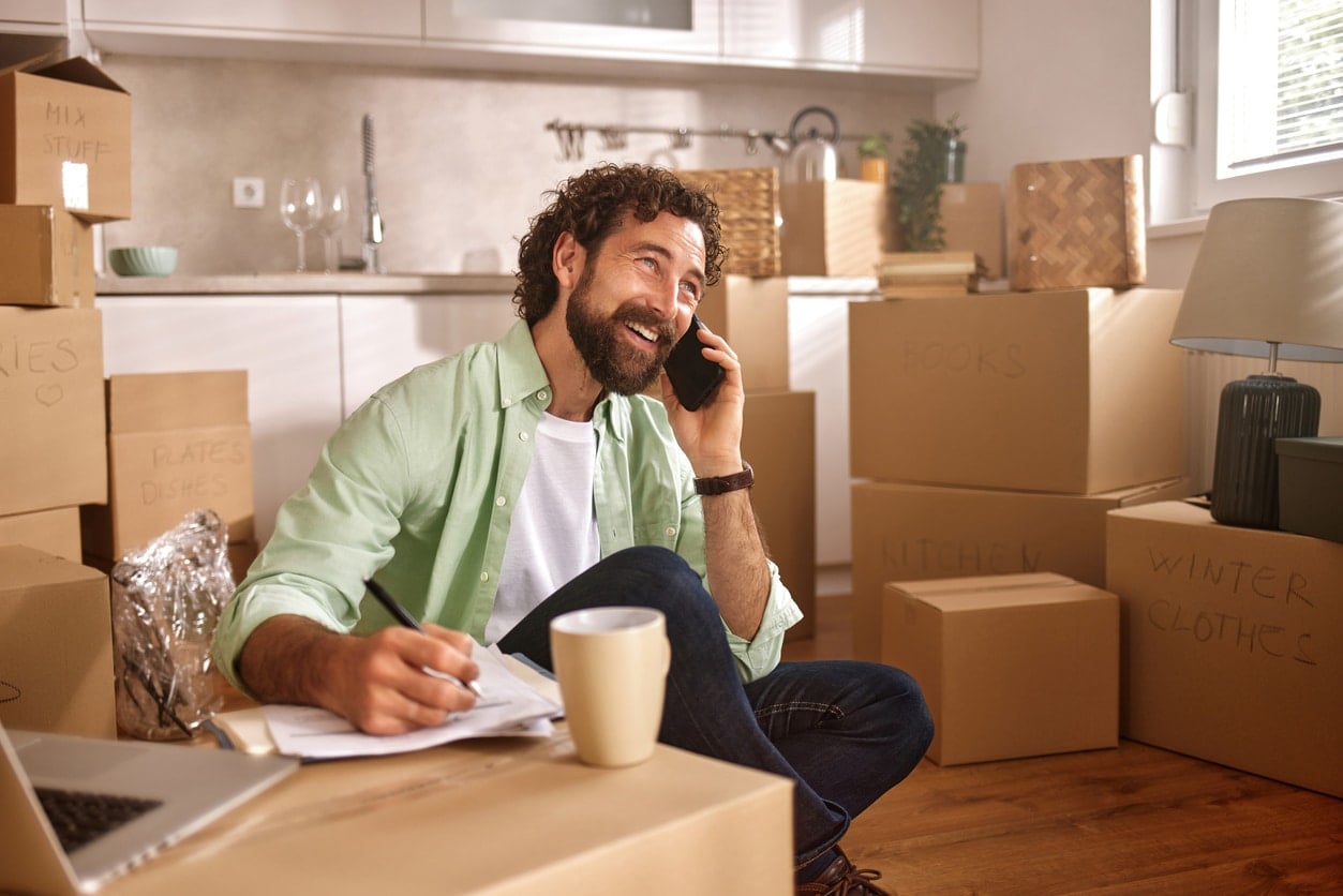 A man talks on a smartphone with a list of questions to ask movers while surrounded by moving boxes