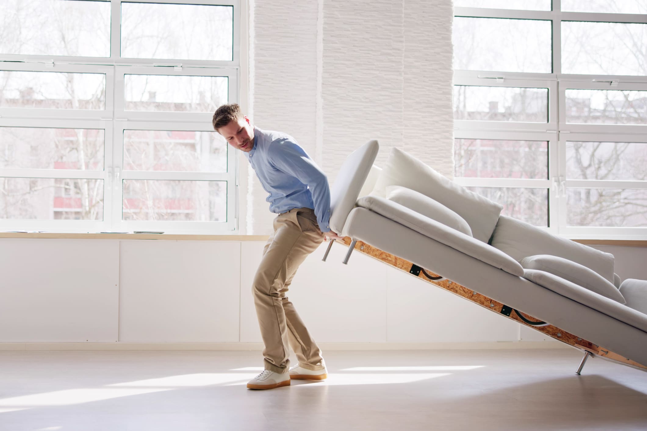 A man in a button-up and slacks moving furniture in a chick all-white city apartment