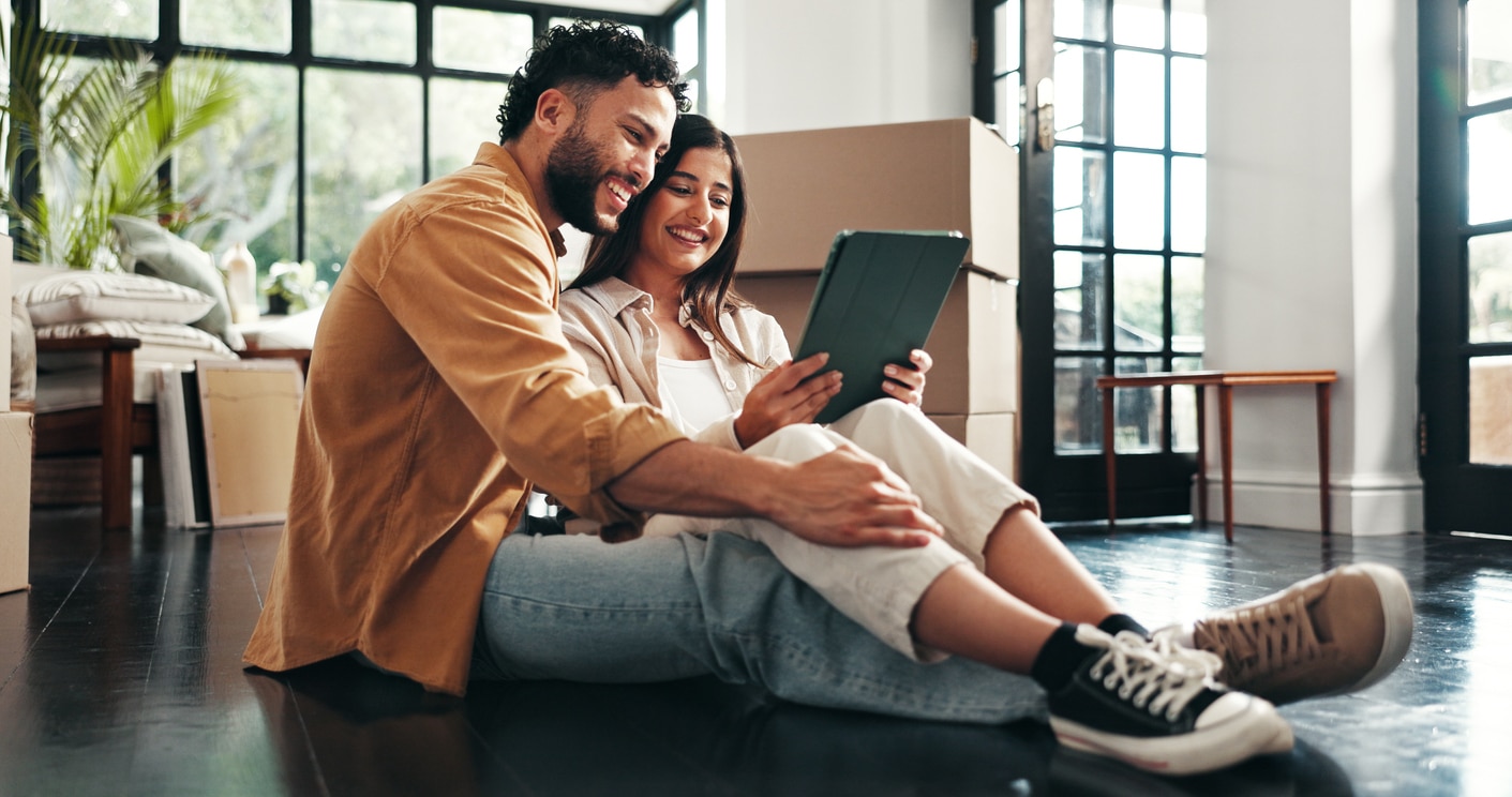 A happy young couple sitting on a living room floor among moving boxes, looking at a tablet together
