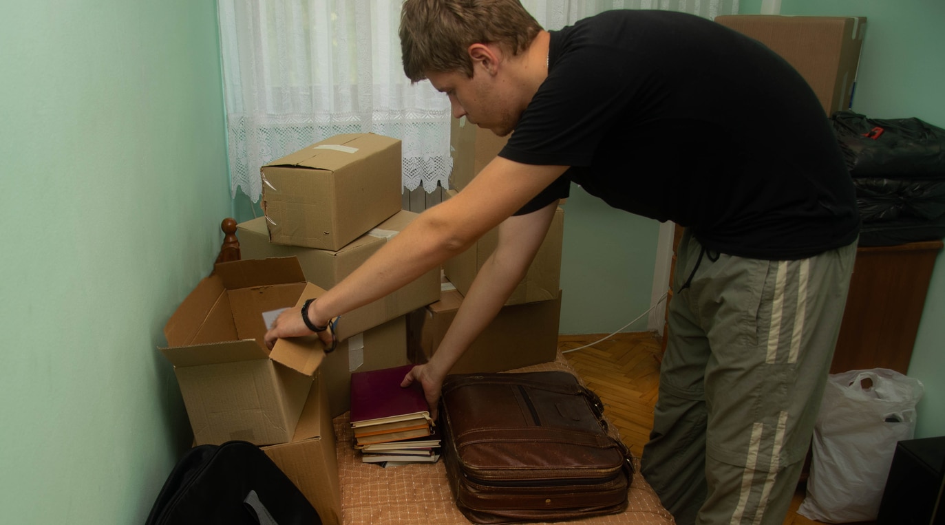 A student in a dorm room bends over to pack or unpack books into a bag among several cardboard boxes