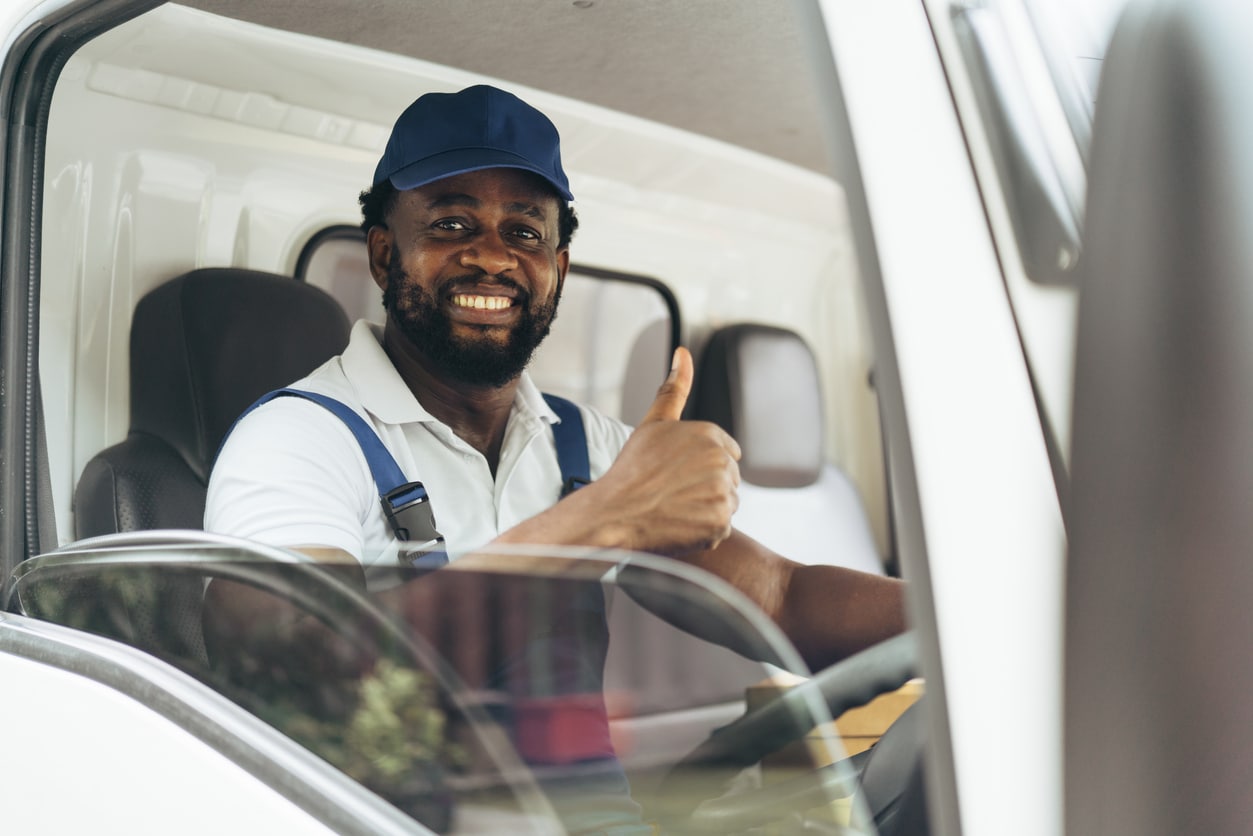 Smiling delivery driver in blue cap and overalls giving a thumbs-up in truck cab
