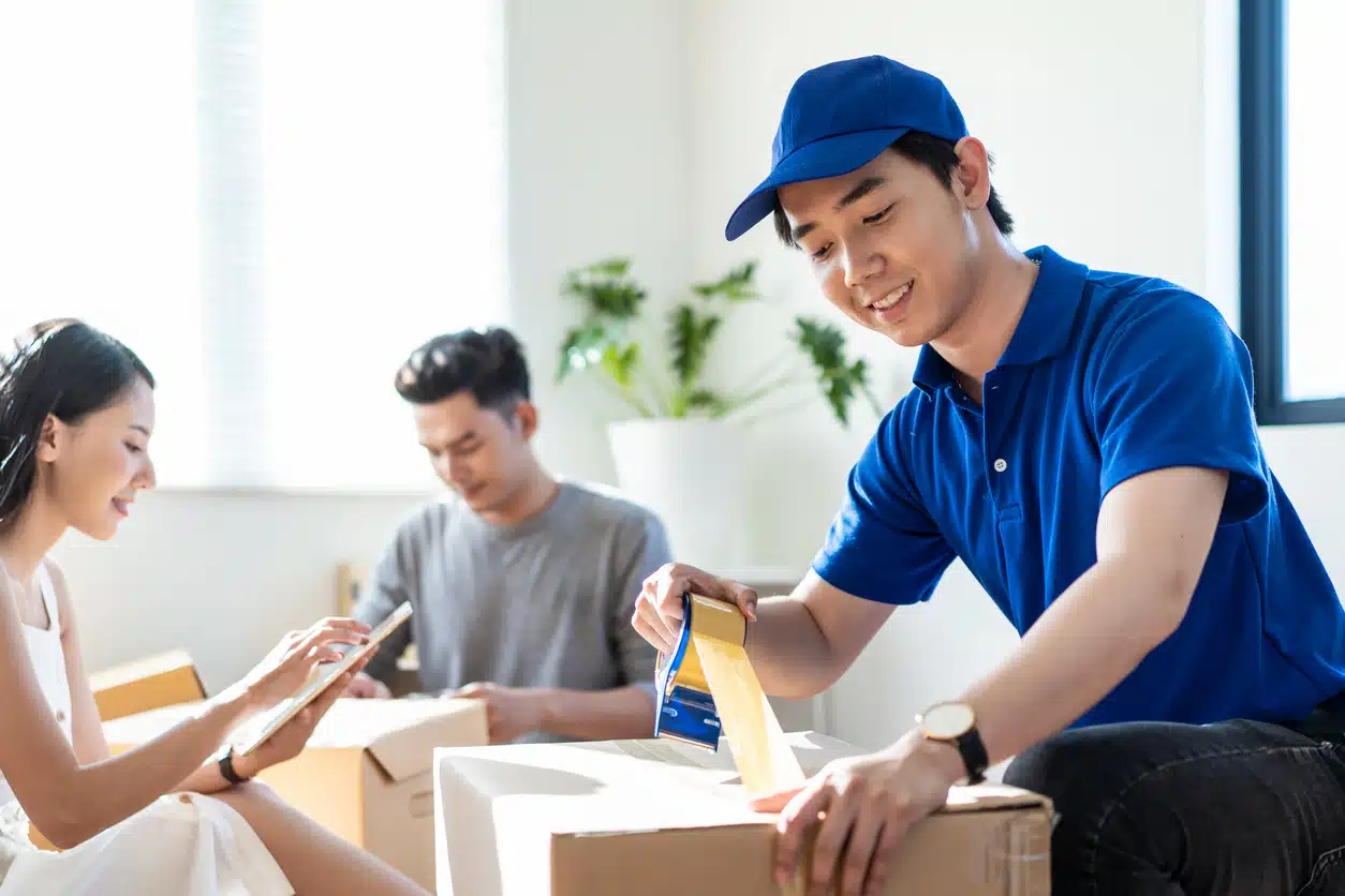 Smiling delivery man uses a tape dispenser to seal a moving box, while a couple work behind him