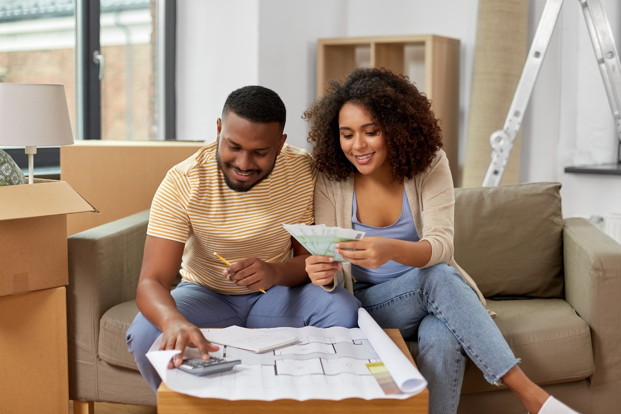 Smiling Black couple on sofa with moving boxes, reviewing blueprints while planning their new home