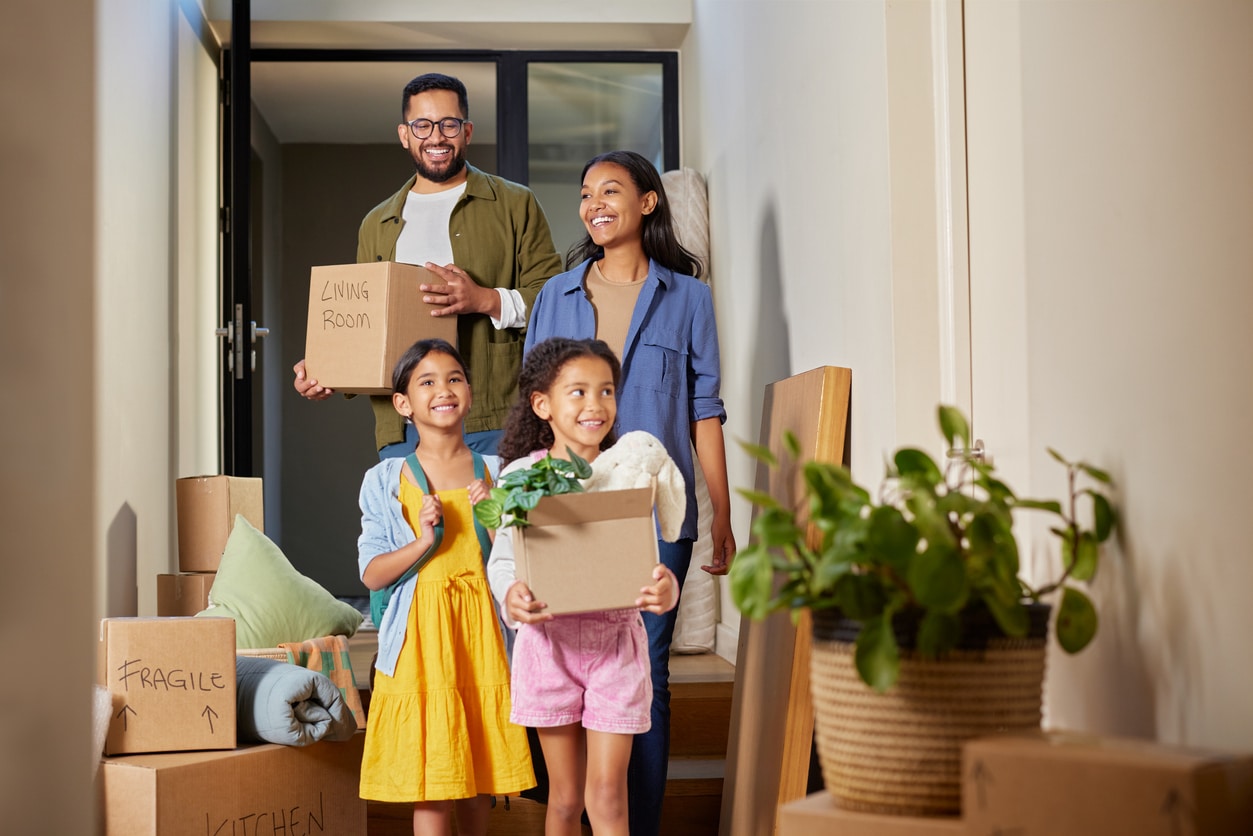 Parents and two young daughters carry moving boxes and a plant inside the entry of their new home