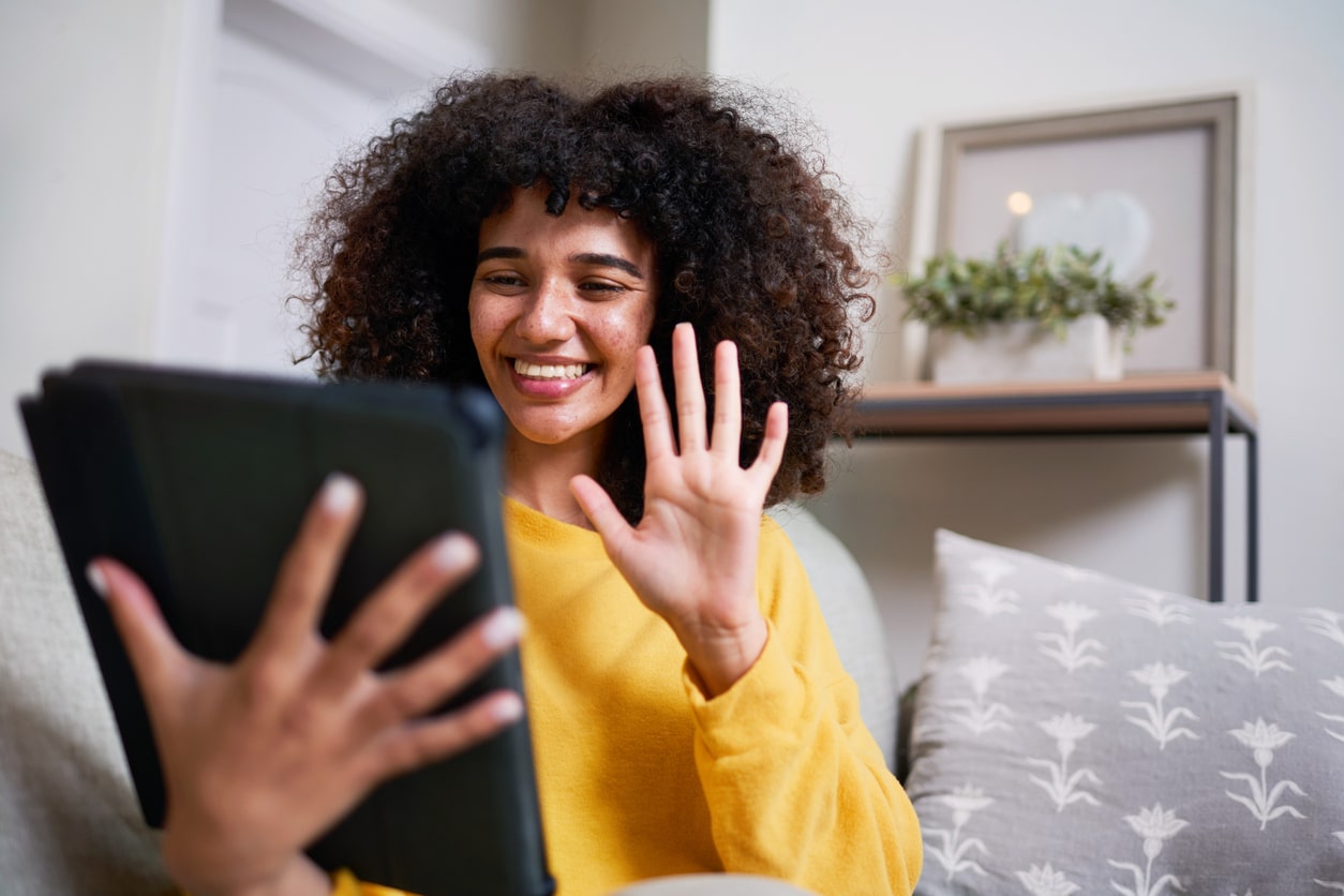 Smiling Woman in Video Call Using Tablet While Seated in Cozy Living Room A young woman in a yellow shirt waves to people on an ipad video call after moving away from friends and family