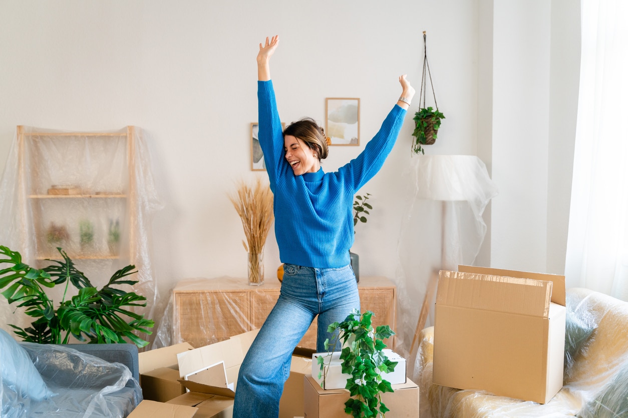 A young woman surrounded by moving boxes in a blue shirt lifts her hands in celebration as she prepares to move away