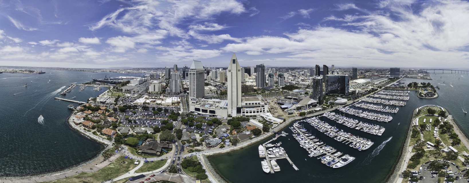 San Diego Aerial Panoramic An aerial view of San Diego’s skyline and bay, featuring downtown buildings, a marina with boats, and a pier under a blue sky
