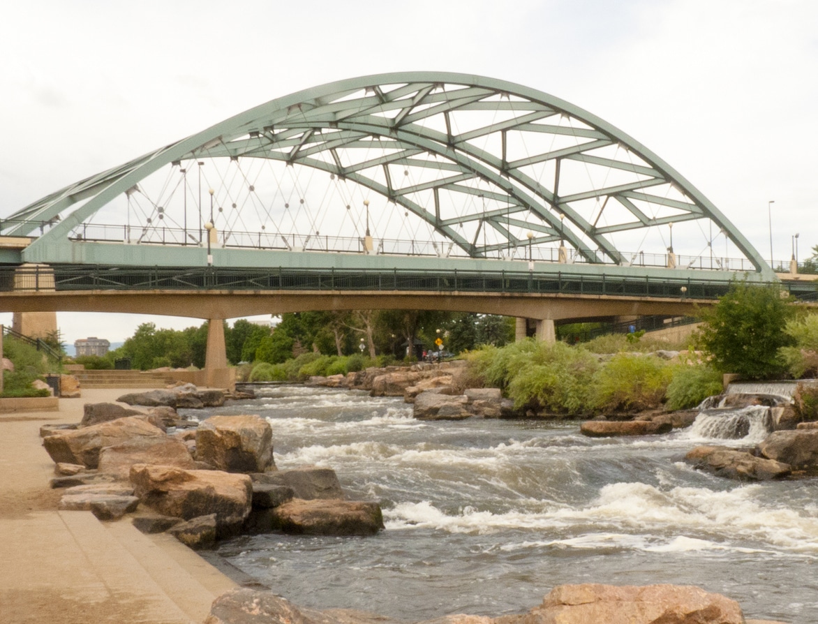 Speer Boulevard Bridge, a teal arch bridge, spans the South Platte River and a water weir in Denver, Colorado