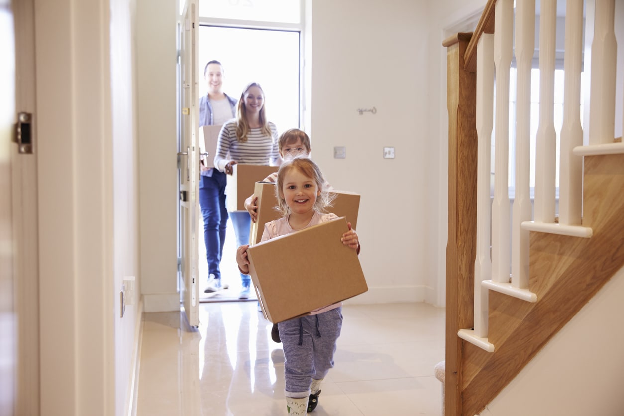 Smiling young girls carry moving boxes into a sunlit new home as their parents get ready to unpack after moving