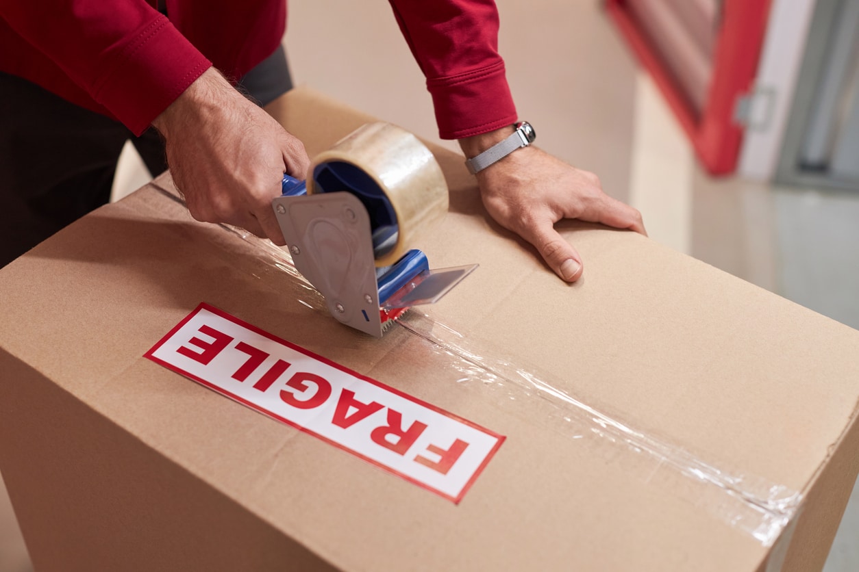 The hands of a person sealing a cardboard box, marked with a label that reads “FRAGILE” in red