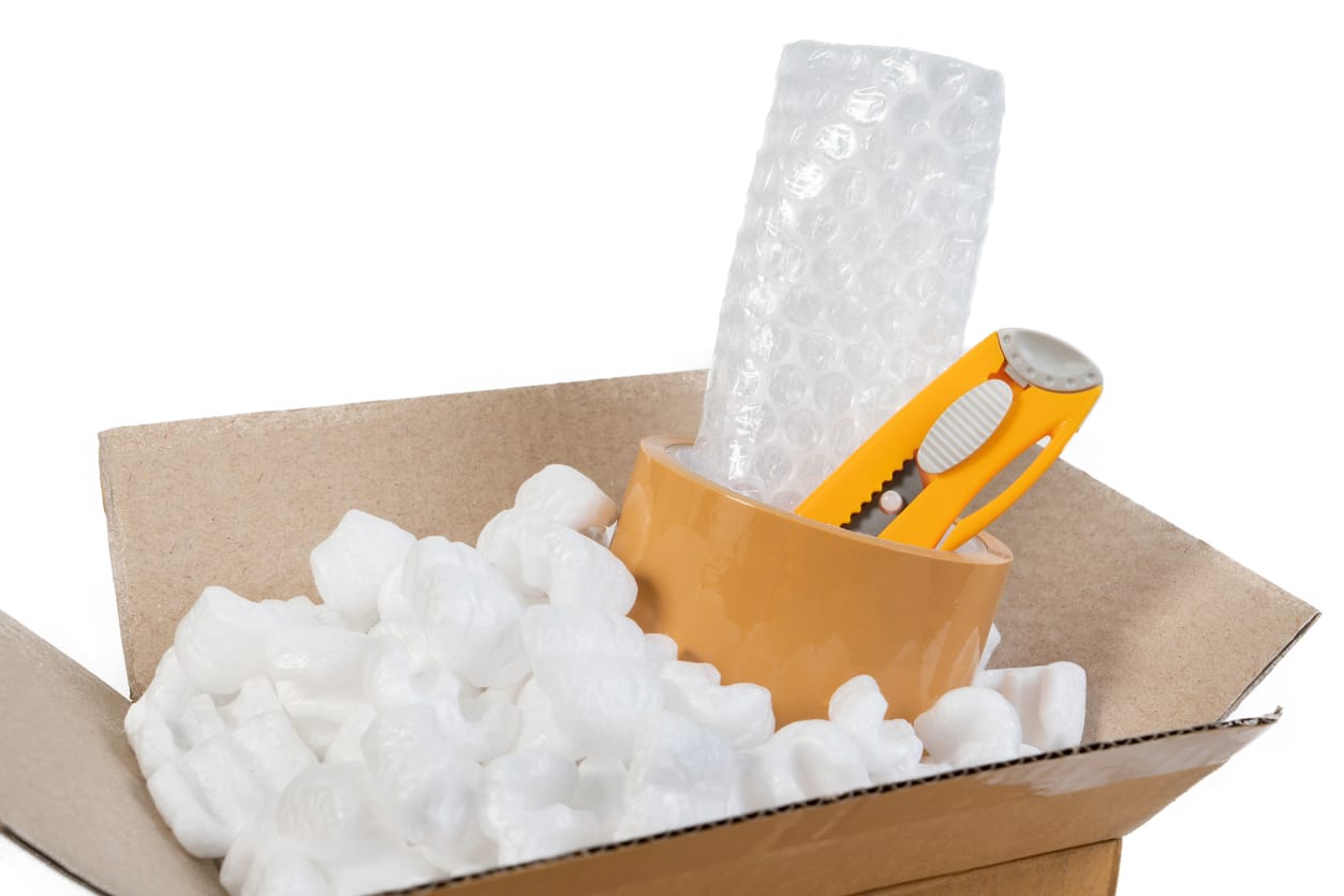 Open cardboard box filled with packing peanuts, bubble wrap, and an orange utility knife against a white background
