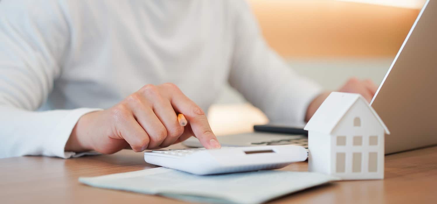 A close-up of a person using a calculator next to a laptop and a small model house on a wooden desk