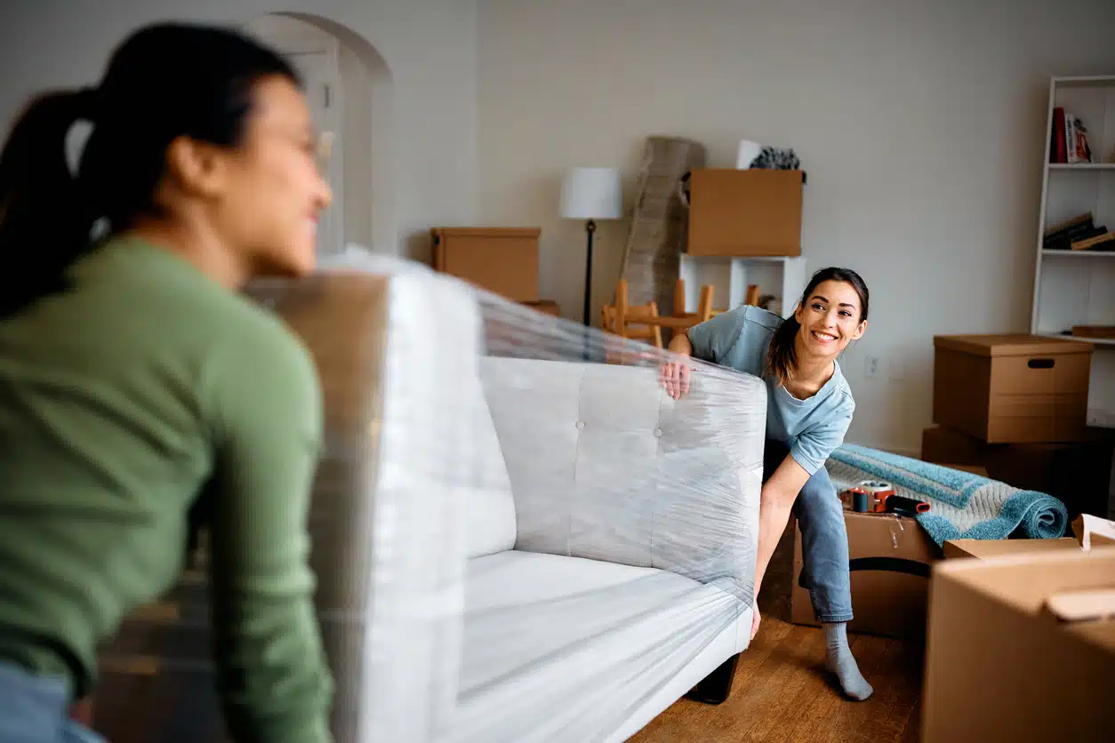 Two women moving a couch while moving, showing the couch wrapped in plastic to prevent damage