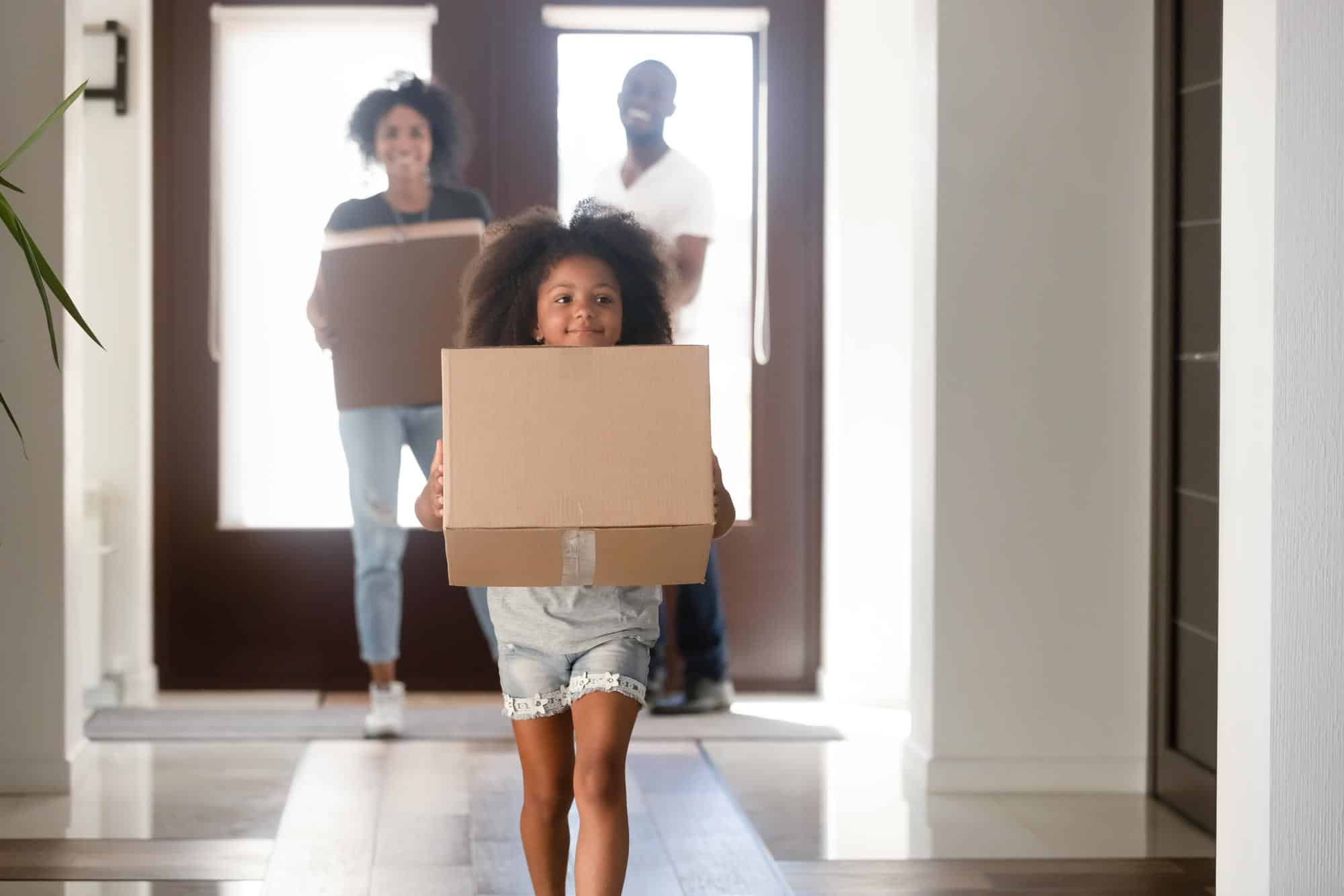 Little girl carrying moving boxes to move cross-country with her parents behind her