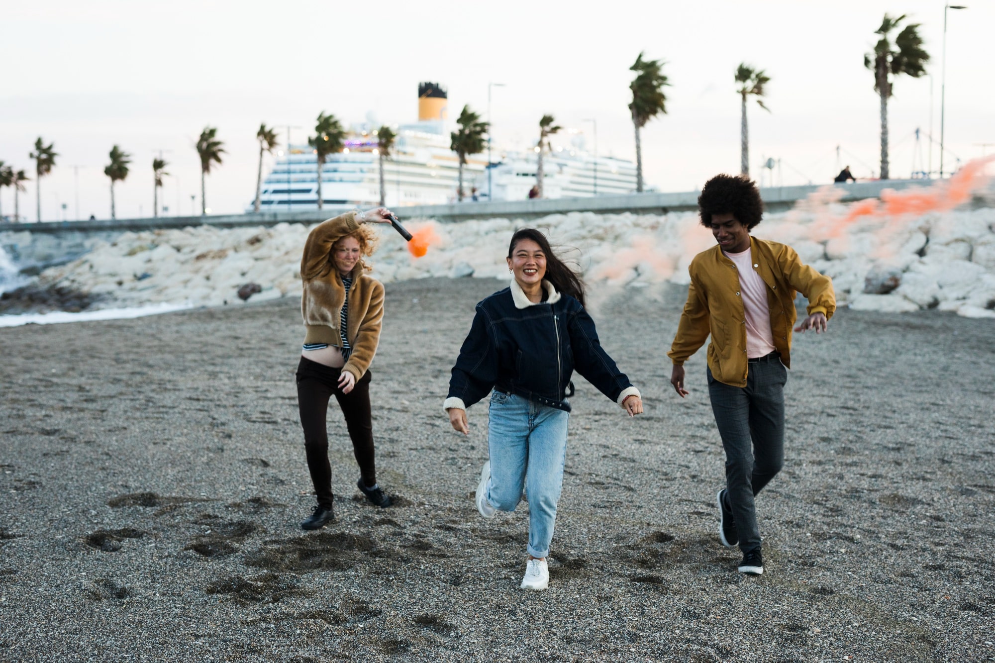 Three friends running on a beach in California after moving to California, one holding a smoke flare, with a cruise ship and palm trees in the background