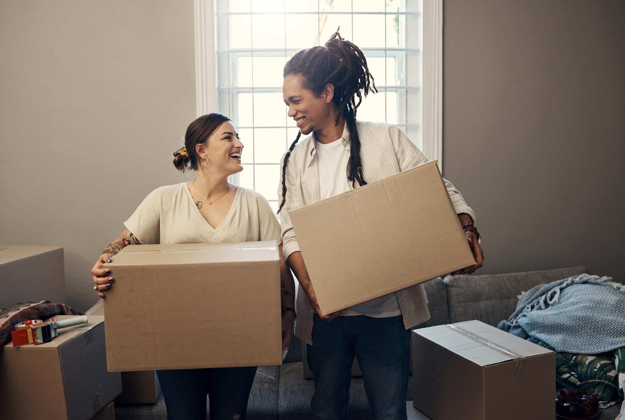 Smiling couple holding cardboard boxes while packing up their home, preparing to move to California