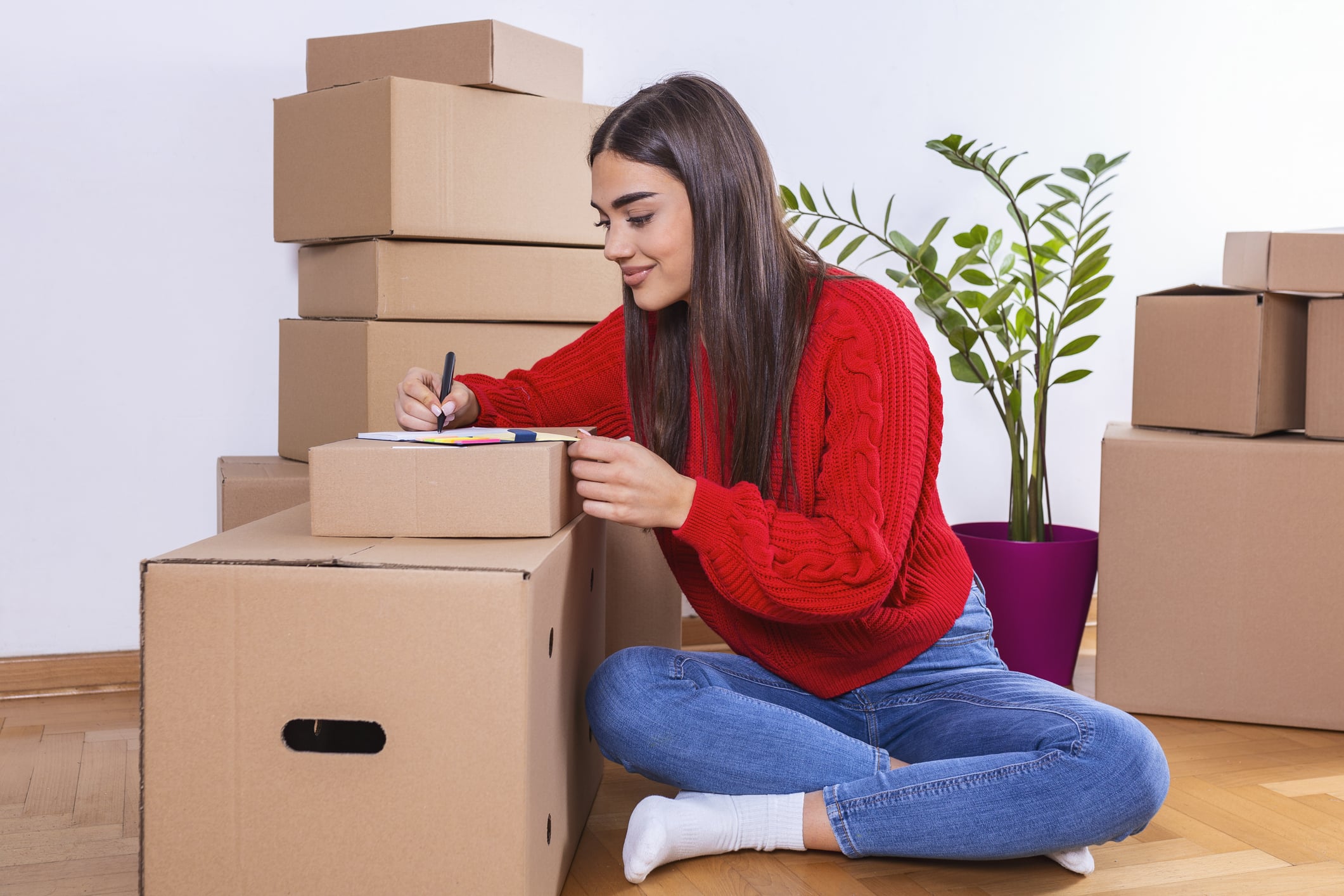 A young beautiful women makes her notes and plans in her new flat,apartment. A woman with long brown hair working on her moving checklist on a pile of boxes and preparing for moving heavy furniture