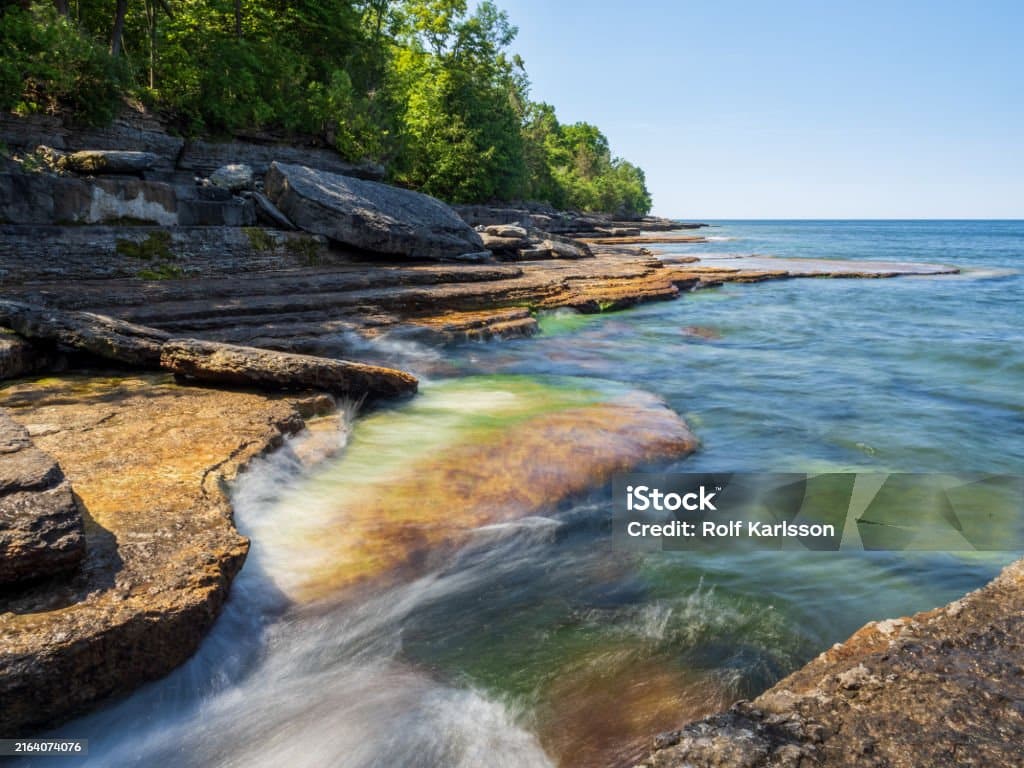 istockphoto-2164074076-1024x1024 Glistening, smooth water cascades over sunlit cliffs draped in moss by Lake Ontario at Robert G. Wehle State Park, upstate New York, framed by a tranquil horizon and a brilliant blue sky.