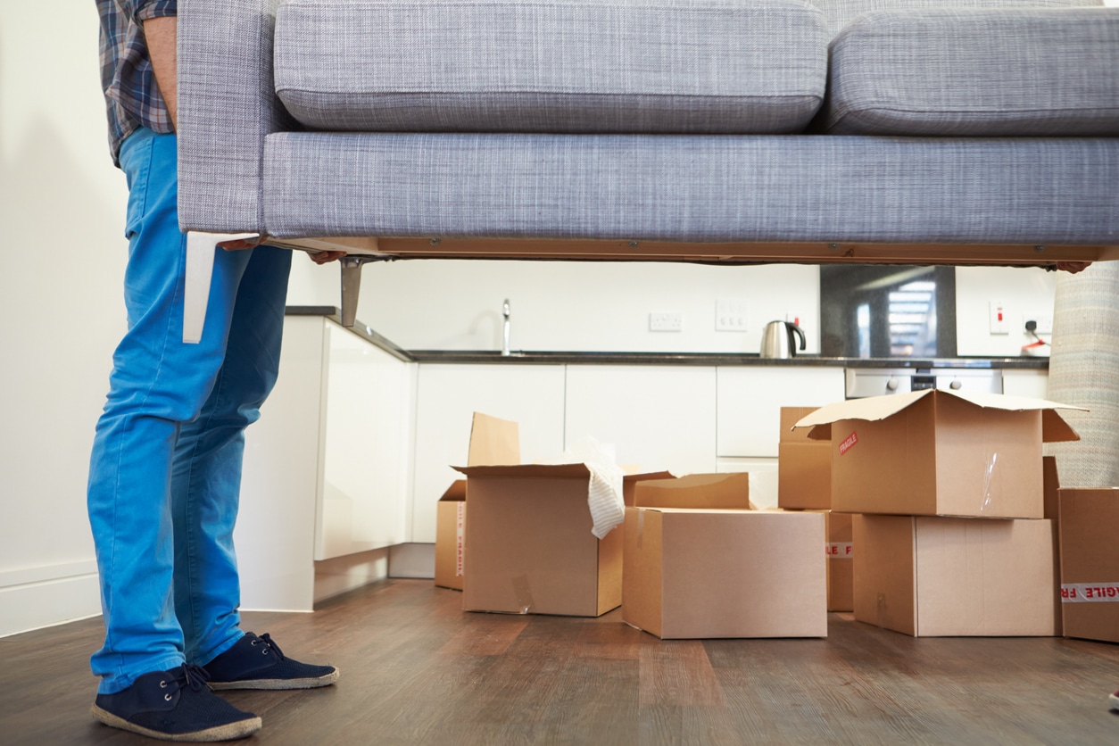Man Carrying Sofa As He Moves Into New Home A person carrying a gray couch, with several moving boxes in the background