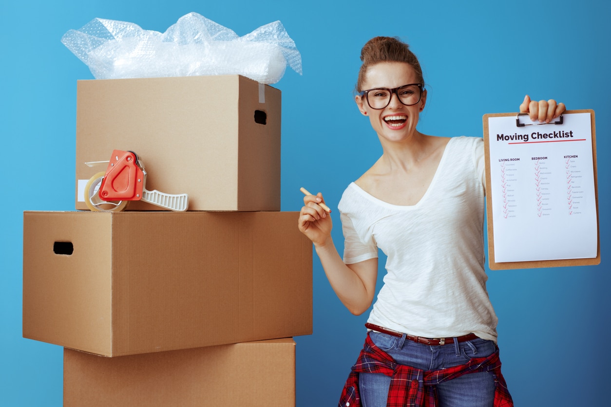 happy woman near cardboard box showing moving checklist on blue A woman happily holding up her moving checklist with several cardboard boxes stacked next to her
