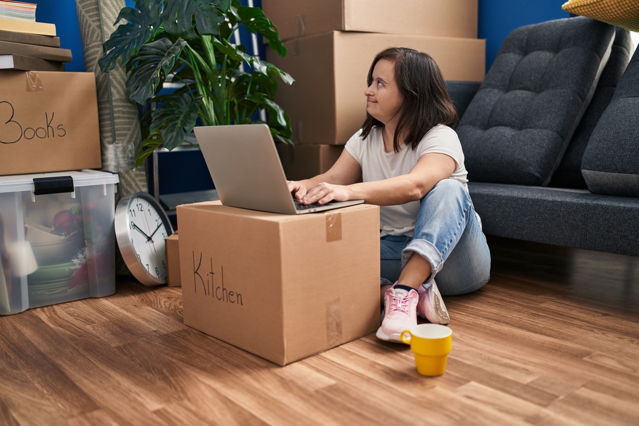 Down syndrome woman smiling confident using laptop at new home A woman utilizing a box labeled “kitchen” as a makeshift desk while sitting on the floor, surrounded by moving boxes