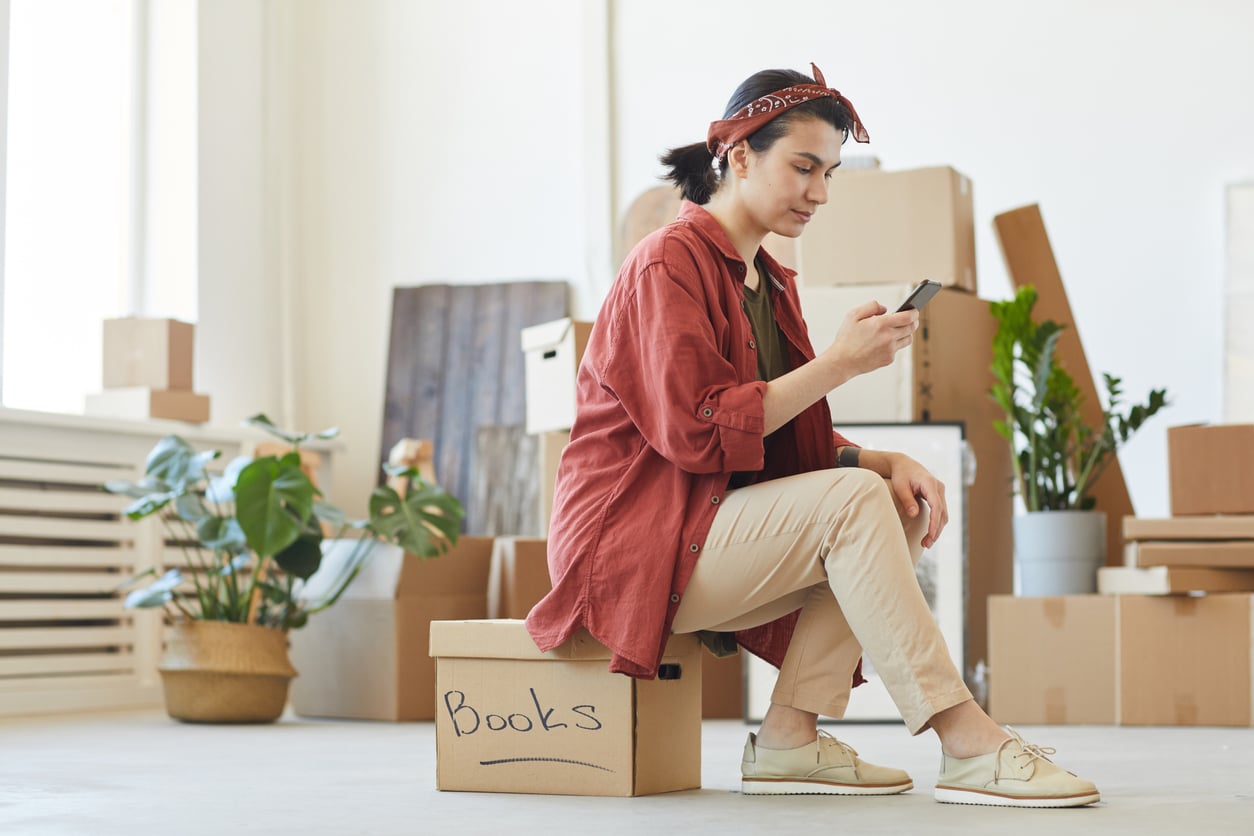 Woman ordering the car A woman looking at her phone on moving day while sitting on a box labeled "Books," surrounded by other boxes