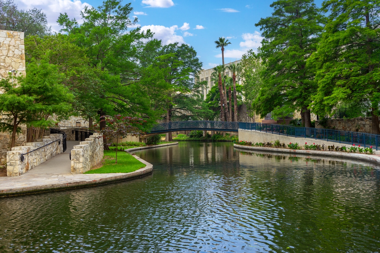 San Antonio River Walk in the daytime, with a beautiful arched bridge, stone walls, and shady trees