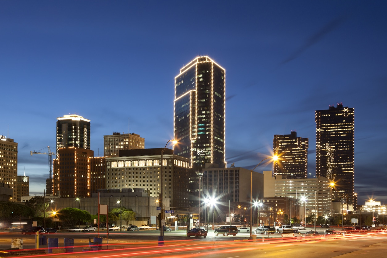 Nighttime cityscape of downtown Fort Worth, Texas, with lit-up skyscrapers and traffic light trails on a busy street