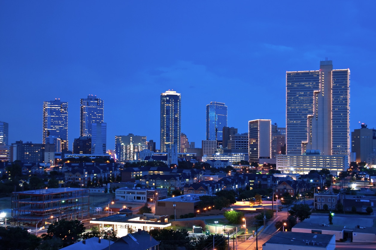 Nighttime cityscape of Fort Worth, Texas, with glowing skyscrapers and city lights beneath a clear, deep blue sky