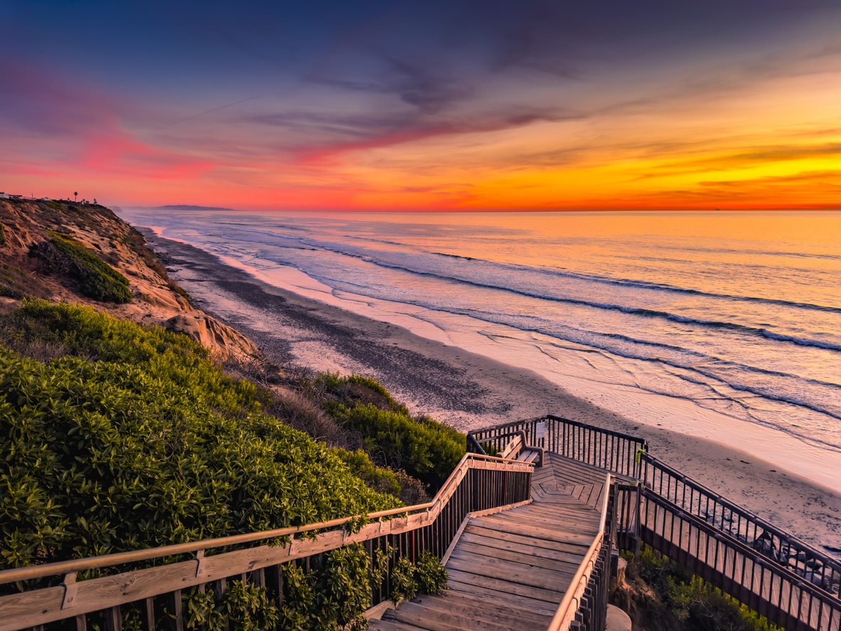 Wooden stairs descend a cliffside to a San Diego neighborhood beach with waves crashing at sunset