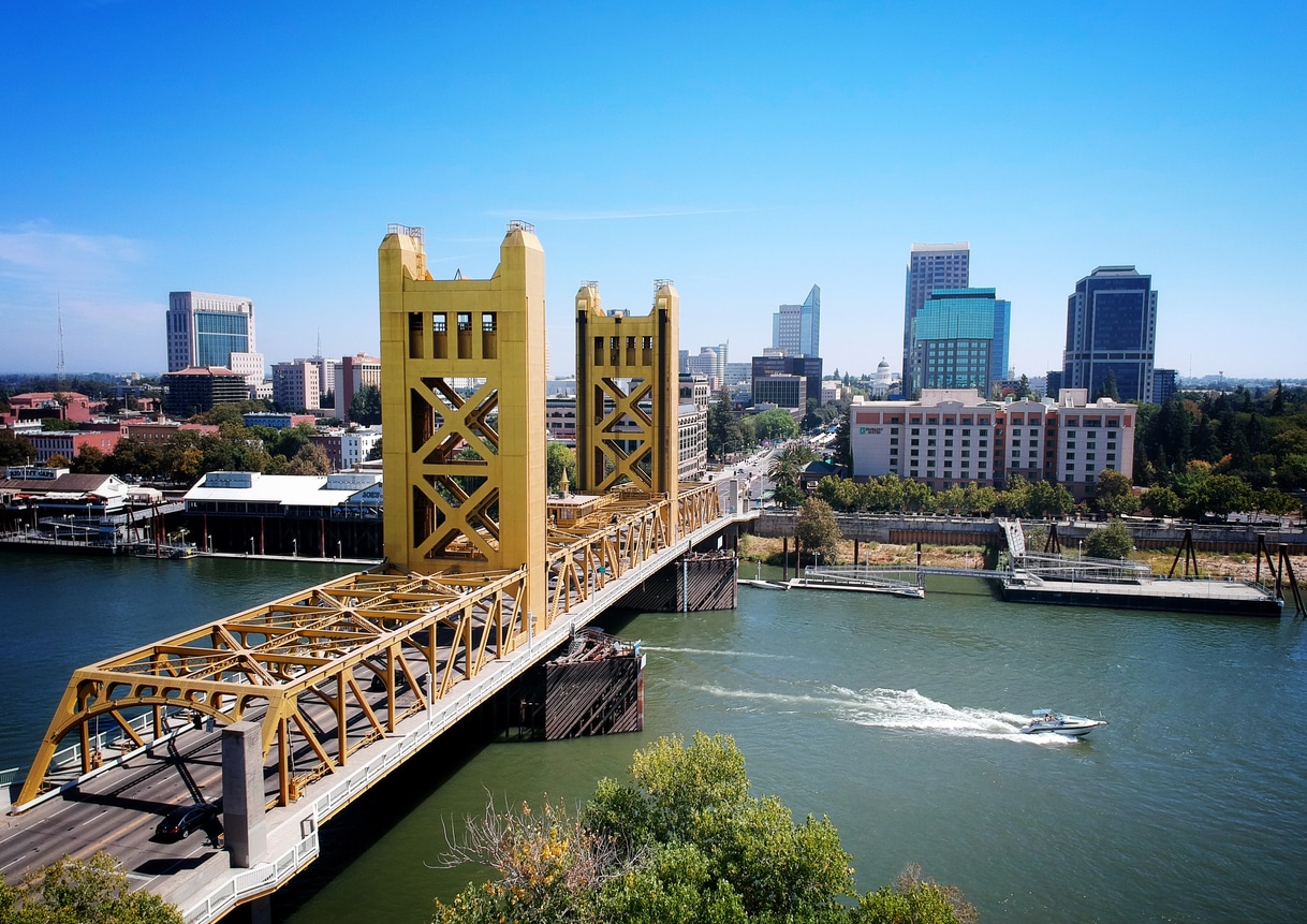 A vibrant blue sky over the Sacramento skyline with modern buildings, a river in the foreground with a small boat