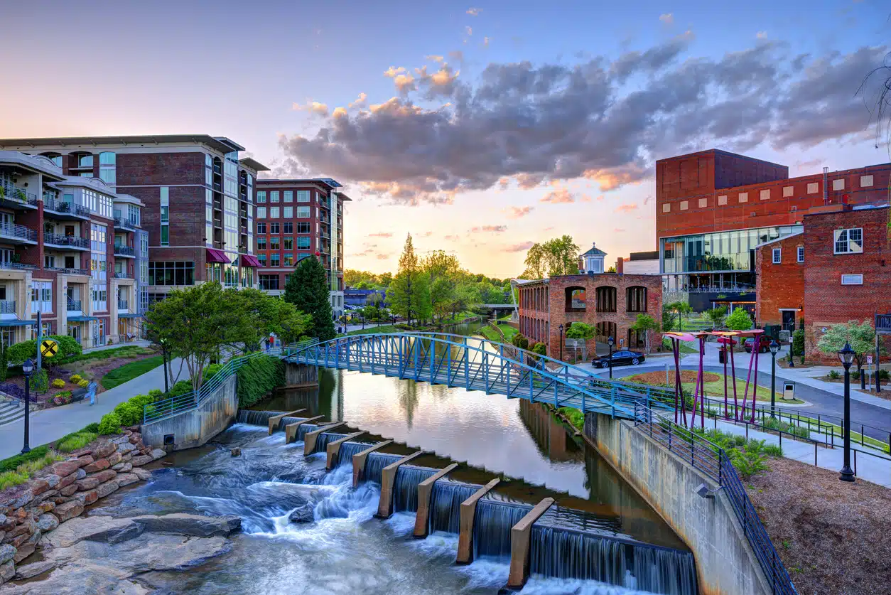 Greenville, SC cityscape at sunset, featuring the Reedy River with a bridge, waterfalls, and surrounding buildings