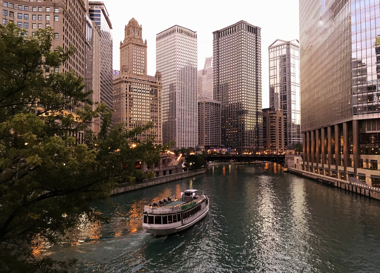 Large Metropolis View With Tall Skyscrapers, City Lights, a Riverboat and Water Reflections Nearing Dusk A panoramic view of the Chicago River and downtown skyline at sunset, a beautiful sight for those living in the Midwest