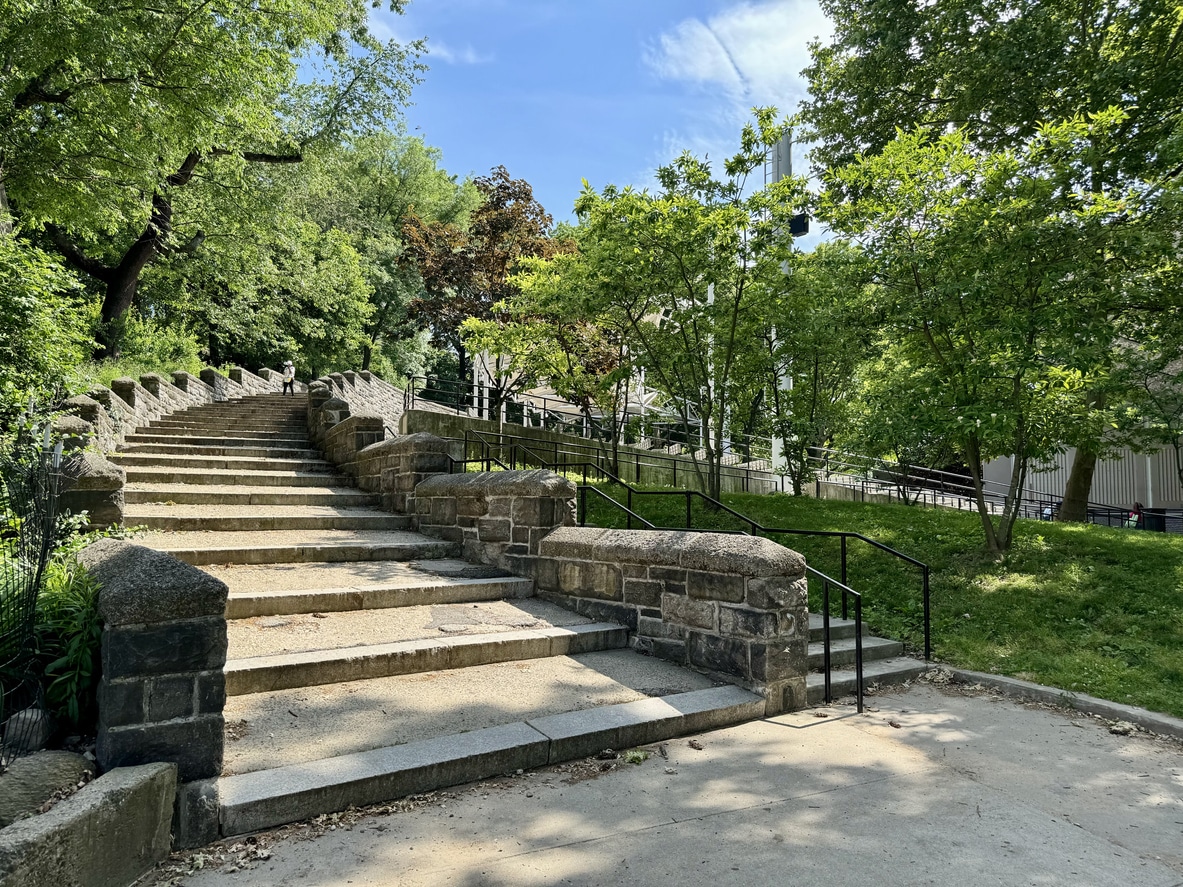 A stone staircase ascends through lush green trees in Harlem's Morningside Park, New York City