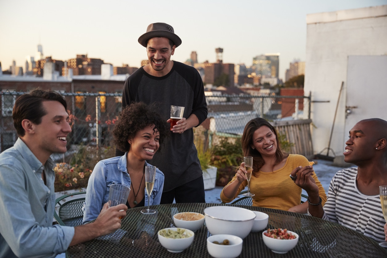 Five friends living in Brooklyn laugh and enjoy drinks and snacks on a sunset rooftop gathering