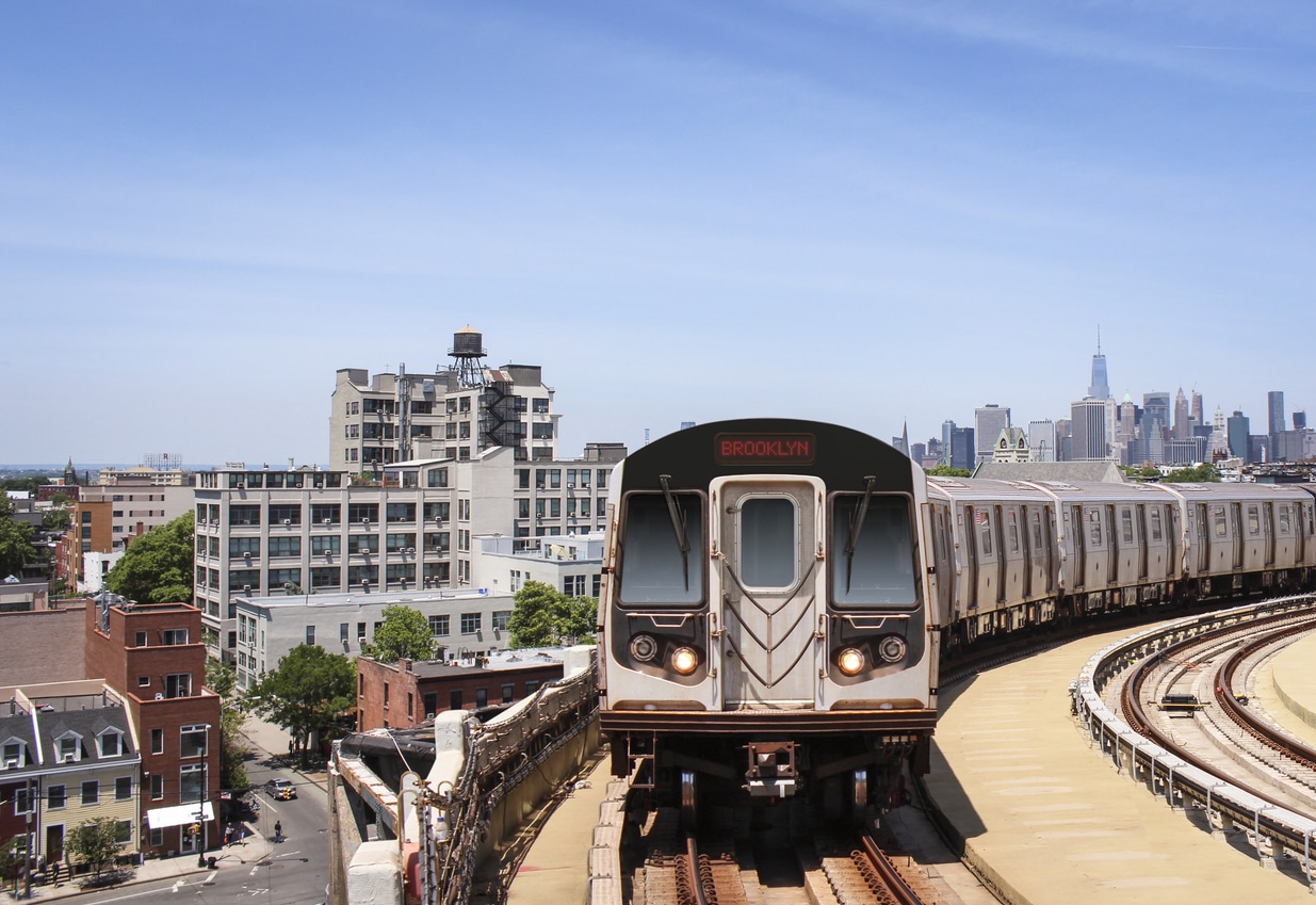 Brooklyn subway train on elevated tracks with city buildings and Manhattan skyline in the distance