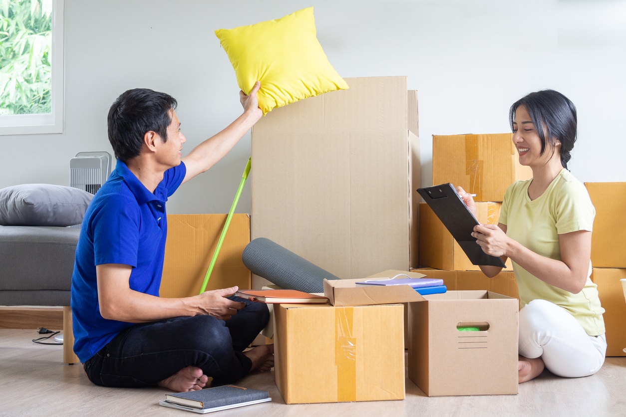 A smiling Asian couple on the floor of a bright room with moving boxes, holding a pillow and a checklist