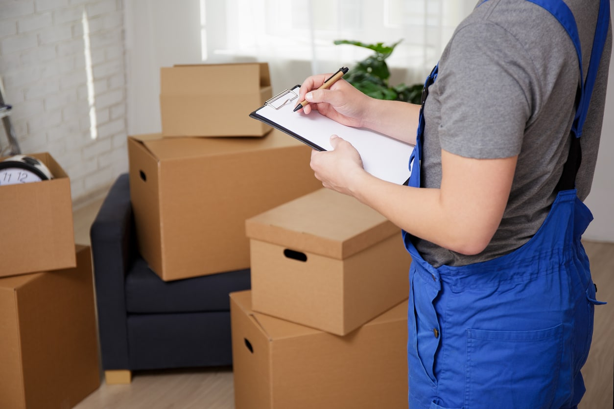 A worker in blue overalls checking inventory on a clipboard amidst cardboard moving boxes in a room