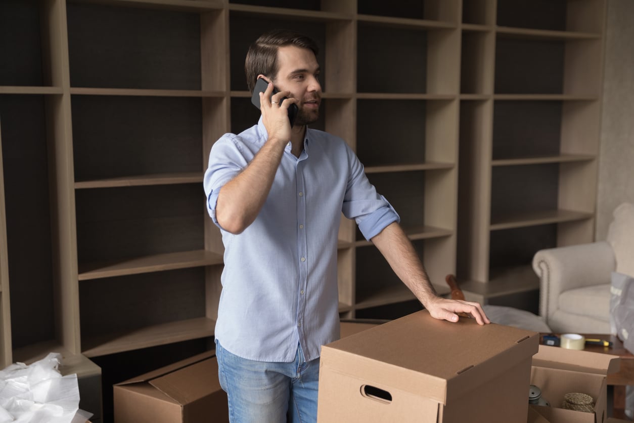 Man in blue shirt talks on phone, standing next to moving boxes and empty shelves in a new room