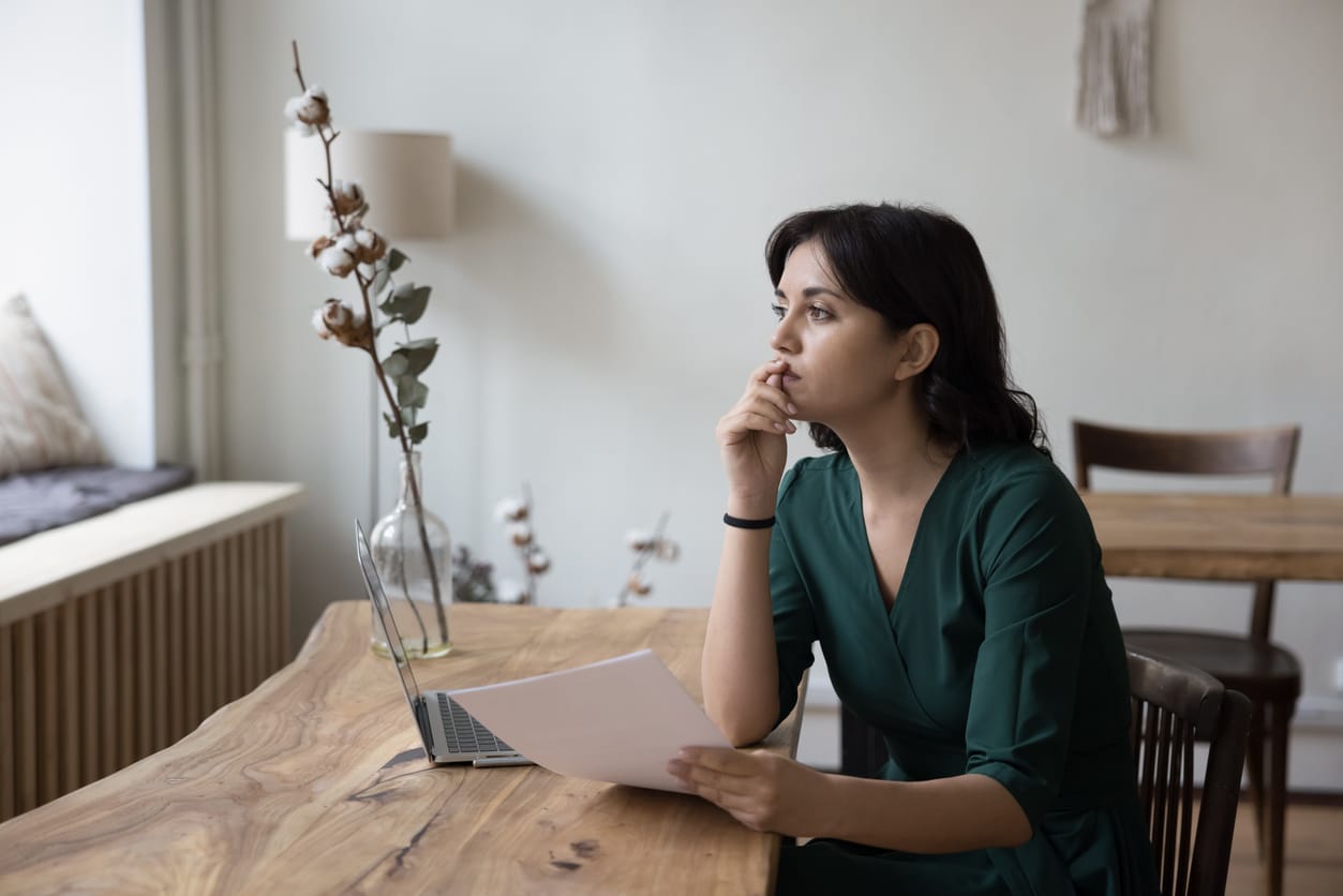 A woman sits at a table with papers and a laptop, looking up thoughtfully in a brightly lit room