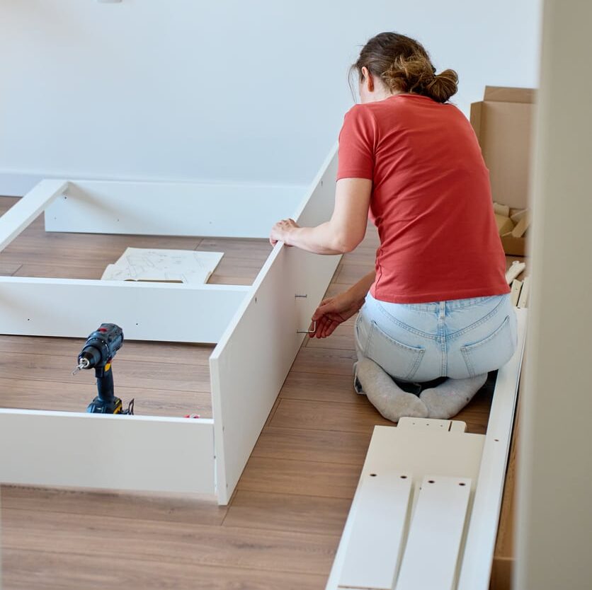 Woman kneels on a wooden floor assembling furniture with a power drill beside her after moving into her new home