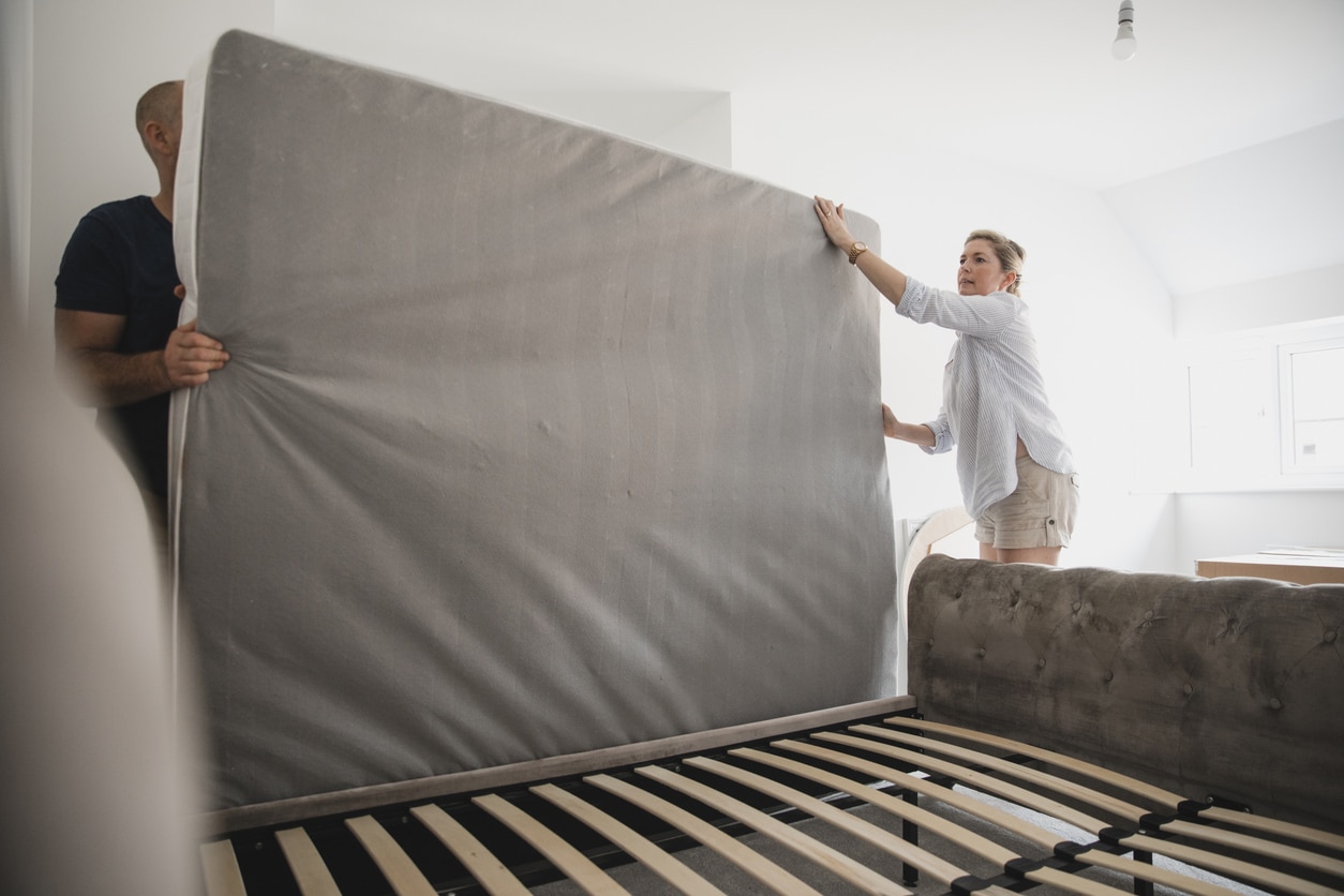 A couple carefully lifts a gray mattress onto a wooden bed frame while setting up their new bedroom after moving