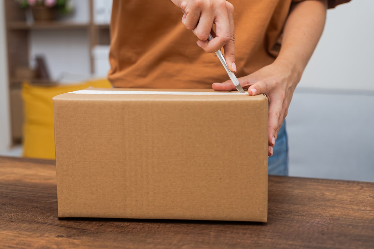 Close-up of a person in a brown shirt using a box cutter to slice open a sealed cardboard moving box
