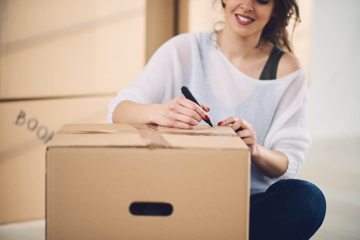 Woman in a white shirt sits down and labels a moving box with a marker as she packs for a move