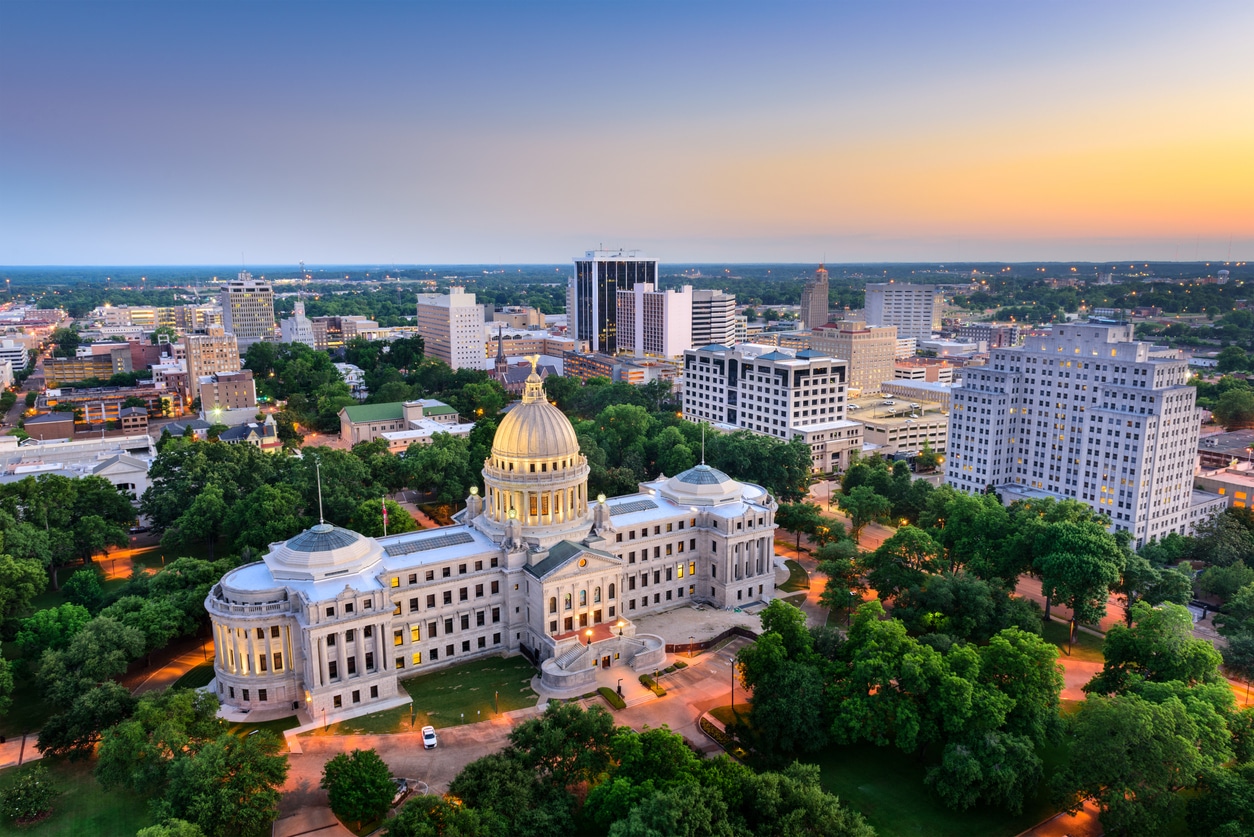 An aerial view captures the Mississippi State Capitol at sunset, surrounded by lush green trees and city buildings