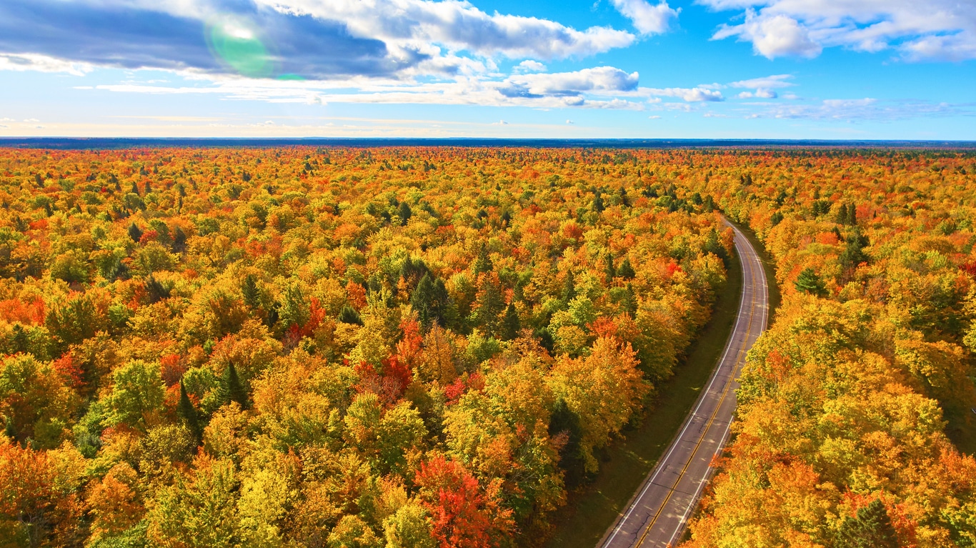 Aerial view of a winding road through a vibrant autumn forest canopy in Michigan under a blue sky with soft clouds